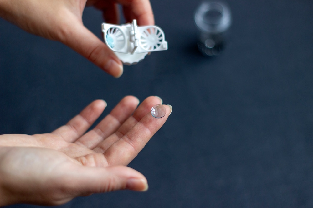 Orthokeratology night lens held on fingertip near contact lens case, representing non-surgical vision correction worn overnight.