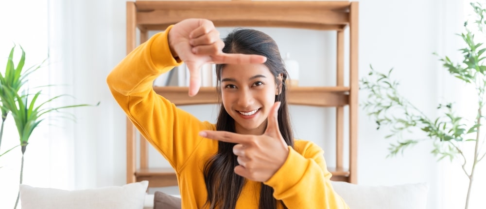 Young woman smiling and making a frame with her fingers in a living room, capturing a happy moment and relaxed home lifestyle.